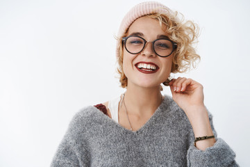 Close-up shot of stylish good-looking young 20s woman with blond short curly hairstyle in glasses and pink beanie wearing warm sweater smiling and laughing broadly looking through glasses at camera