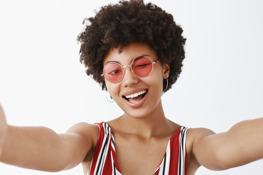 Hey, what is up. Playful hot African American with afro hairstyle, pulling hands towards camera to make selfie, winking joyfully and smiling broadly, making new profile pic for social network