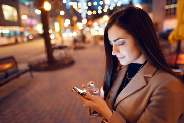 Fototapeta premium Beautiful girl in a evening city. Stylish brunette in a brown coat. Woman in a Chicago. Lady with a phone