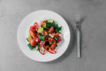 Top view of fresh vegetable salad prepared of red pepper, radish, tomatoes, cucumbers and parsley in white bowl, fork near. Vegetarian dish concept. Healthy nutritious spring salad