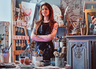 Beautiful painter in her own studio. She is standing with crossed hands. There are a lot of artist's tools at background.