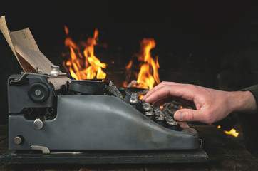 A writer is typing a text on a typewriter on his desk on a burning fire in fireplace background.