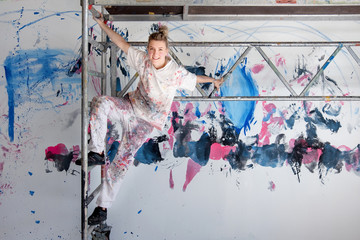 young pretty woman painter hangs on mobile scaffolding against colorful painted wall.
