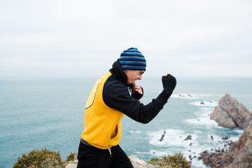Adult bearded man in sportswear practicing punches during kickboxing workout on rocky cliff near sea
