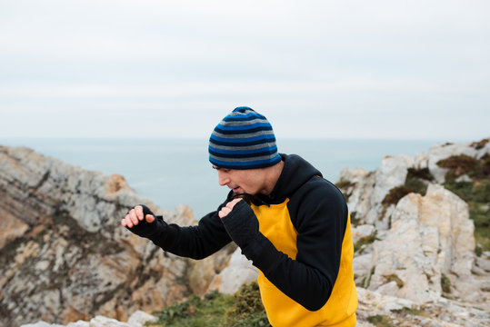 Adult bearded man in sportswear practicing punches during kickboxing workout on rocky cliff near sea - Powered by Adobe