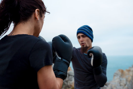 Man and woman in boxing gloves punching each other while standing on cliff against sea and cloudless sky