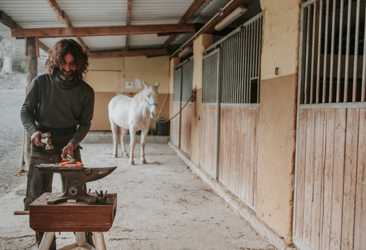 Adult farrier using hammer and tongs to forge hot horseshoe on portable anvil near stable on ranch