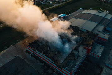 Fototapeta premium Aerial view of burnt industrial warehouse or logistics center building after big fire with huge smoke from burned roof, drone shot