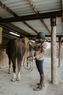 Side view of young female in helmet putting saddle on wonderful horse near stable on ranch