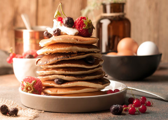 side view of tasty traditional american pancakes in stack with sour cream, fresh berries on white plate near bowl with raw brown eggs and glass bottle on table