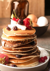 stack of delicious pancakes with sour cream and fresh berries on white plate near bowl with raw brown eggs and glass bottle on table