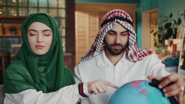 Close up view of arabic couple deciding where to travel, young attractive man holds and rolls the globe when the woman points with her finger on the continent, pleased with the results. Travel concept