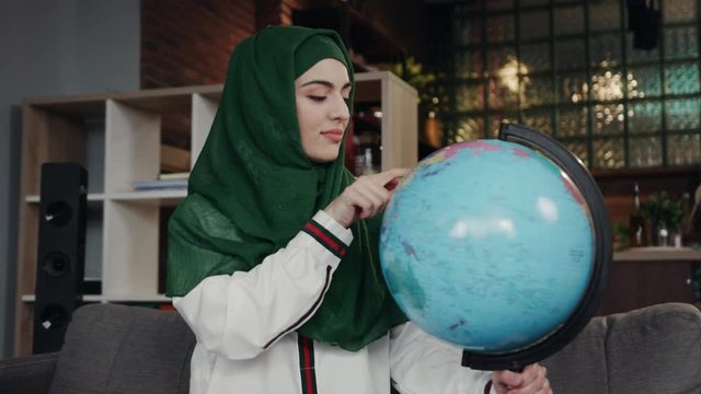 Close up portrait of Arab woman looking at globe, whirling it and pointing at the continent, deciding where to go, satisfied with result happily dances. Planning, vacation, having fun, travelling