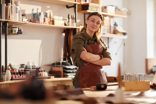 Warm toned portrait of smiling female artisan standing with arms crossed while posing in workshop