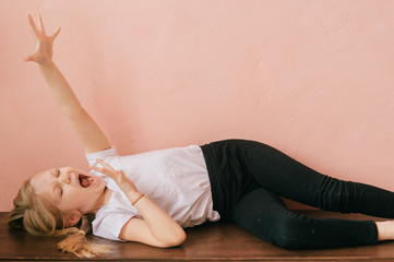 Obraz premium Lifestyle portrait of clowning little girl make faces and shouting on pink wall background. Funny expressive female with ridiculous face kid lying on table and screaming like crazy with gesturing hand