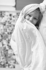 Vetrtically turned black white portrait of little girl lying on bed at home under white blanket and looking ar camera with decorative wall on background.