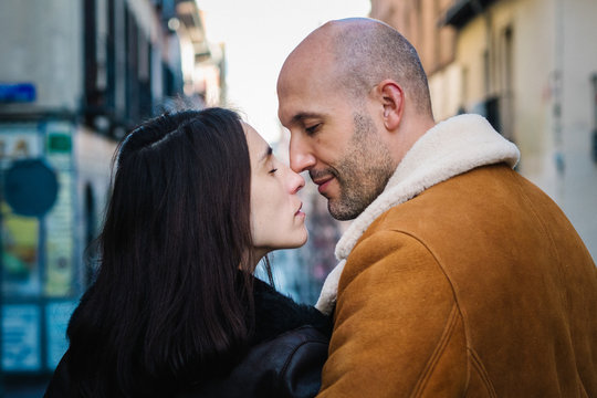 Side view of bald adult man embracing and kissing pregnant wife while standing on narrow city street together