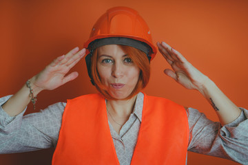 Adult female with red hair in working uniform on orange background. Portrait of woman in bright orange protective hardhat and vest looking at camera on orange background