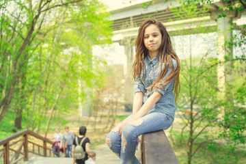 Glad girl sitting on railing Pleased girl in casual outfit looking at camera while sitting on wooden railing of stairway in green park