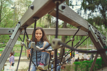 Obraz premium Cute girl having fan on playground. Charming little brunette girl in jeans suit hanging on grid and looking at camera on playground in park