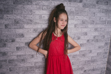 Children model in a red dress against a brick wall. A little girl with long hair standing near a gray brick wall