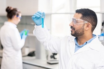 Obraz premium Content satisfied handsome middle-eastern modern chemist in white coat looking at test tube while checking color of liquid in laboratory