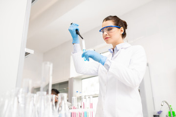 Content experienced young lady chemist in lab coat using dropper and test tube while mixing...