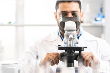 Concentrated busy young Arabian biological scientist sitting at desk and looking in microscope...