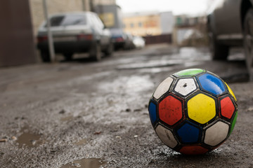 Fototapeta na wymiar children's soccer ball game is on a dirty broken street road among puddles and residential yards of houses, near parked cars