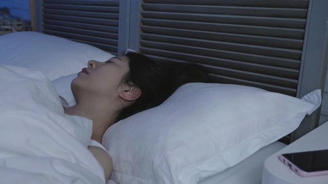 Sleeping asian woman turns head around while dreaming in double bed in dark bedroom. side view young lady lying in comfort white soft pillow frowning having nightmare in midnight. bad sleep quality