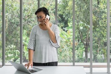 Asian woman talking on her mobile phon while using laptop in front of window