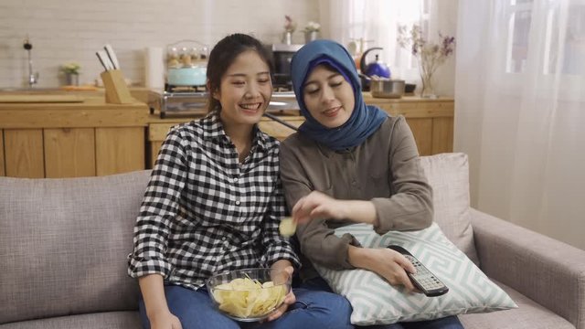 happy multi female best friends sit on couch watch funny film on TV. group of asian arabic women eating snacks laughing talking with comedy program on television holding controller at home kitchen
