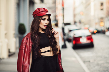 Beautiful fashionable sexy woman with makeup posing on city street wearing a red jacket, hat and sunglasses.