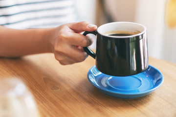 Closeup image of a woman's hand holding a black cup of hot coffee on wooden table