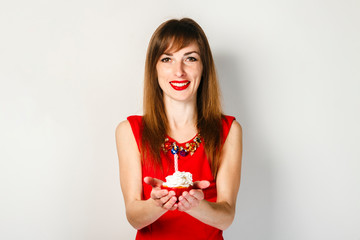 A young girl in a red dress is holding a cupcake with candles on a light background. Birthday celebration concept.