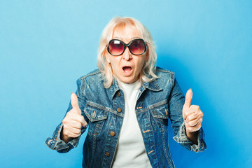 An old lady in a denim jacket and sunglasses is showing a gesture with her hands. Thumb up on a blue background. Concept fashionable grandma, old woman, funky action.