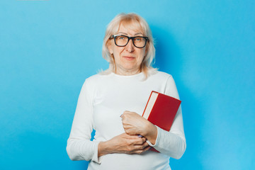 An old woman in glasses with a happy face holding a book in her hands against a blue background. Concept old lady reads books, education, book club.