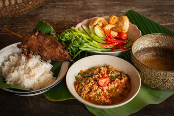 Thai Northern Style Pork and Tomato Chili Relish, nam prik ong in white bowl on wood table there are side dishes of fresh vegetables, cooked rice and  water bowl placed around.