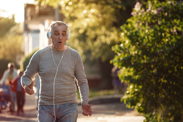 Senior man is walking along the sidewalk listening to music and singing a song