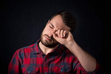 young man in plaid shirt rubs his eyes against black background