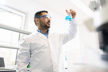 Serious concentrated young middle eastern pharmacist in lab coat and safety goggles standing in laboratory and working on medication