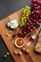 Cheese head, bunch of grapes, honey, nuts and wineglass on wooden board and black background