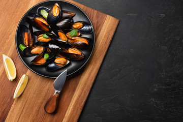 Seafood mussels and basil leaves in a black plate with wineglass, lemon, knife on wooden board and stone table