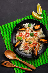 Fried rice with seafood mussels, shrimps, basil in a black plate with wooden spoon and fork on green bamboo mat and stone table