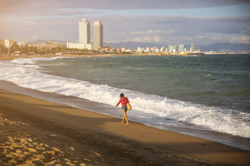 Girl with a bag walking on the beach on sunny day. Stylish hipster near the waves on the sea. Holiday travel concept. Barcelona Spain.