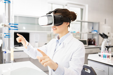 Smiling smart young female scientific researcher gesturing hands while using virtual reality simulator in laboratory