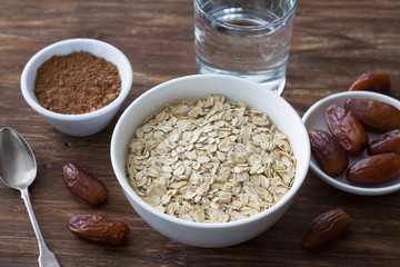 Raw oatmeal in a white bowl, dates fruits, cocoa and a glass of water, ingredients for delicious healthy breakfast on a wooden background