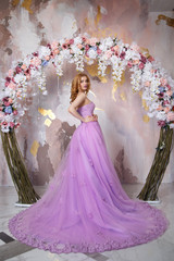 The beautiful girl the blonde in a smart long lilac dress with a train costs near the decorative arch from flowers.