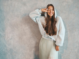 Young beautiful smiling woman looking at camera. Trendy girl in casual summer hoodie and skirt clothes. Funny and positive female posing near gray wall in studio. Showing peace sign