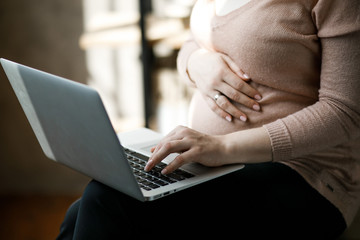 Naklejka premium Pregnant woman working on laptop. Cropped image of pregnant businesswoman typing something on laptop while sitting at her working place in office